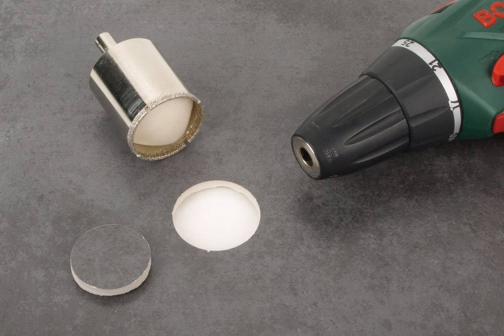 Perceuse avec un adaptateur de scie-cloche à côté d'un trou foré sur une surface grise.