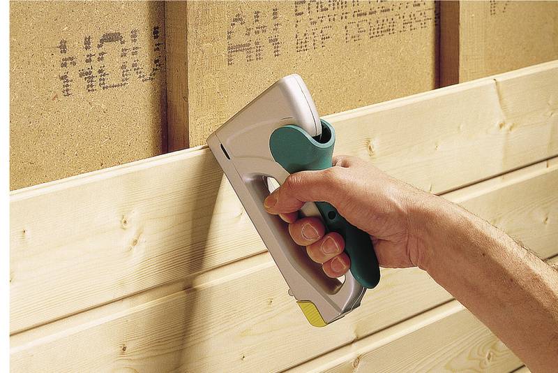 Une main tient un pistolet à agrafes et fixe des panneaux de bois sur un mur.