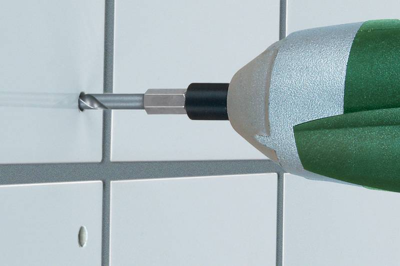 Un foret traverse un carreau blanc selon un motif quadrillé. Le foret a une poignée verte.