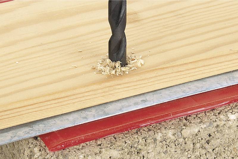 Un foret perfore une planche de bois posée sur une surface en béton. Les copeaux autour du trou indiquent un perçage en cours.