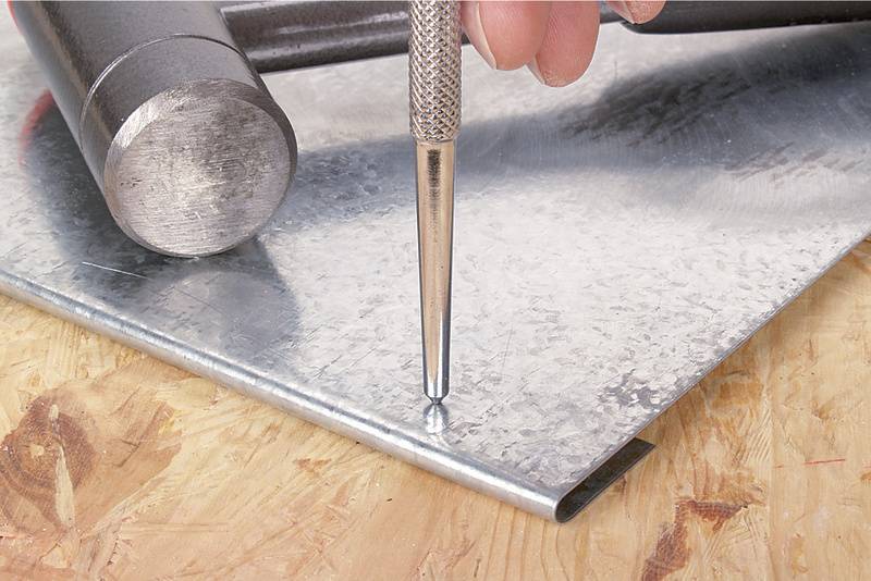 Une main tient un pointeau sur une plaque métallique, prête à poinçonner. Un marteau est posé à côté sur la surface en bois.