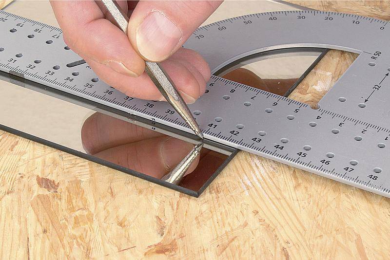 Une main coupe du verre avec un coupe-verre le long d'une règle métallique posée sur une table en bois.