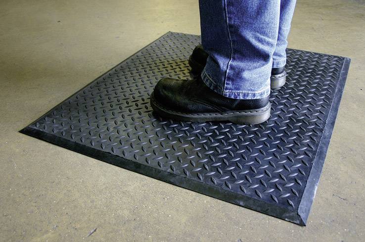 Une personne se tient debout sur un tapis noir antidérapant posé sur un sol en béton, portant un jean et des chaussures de travail noires.