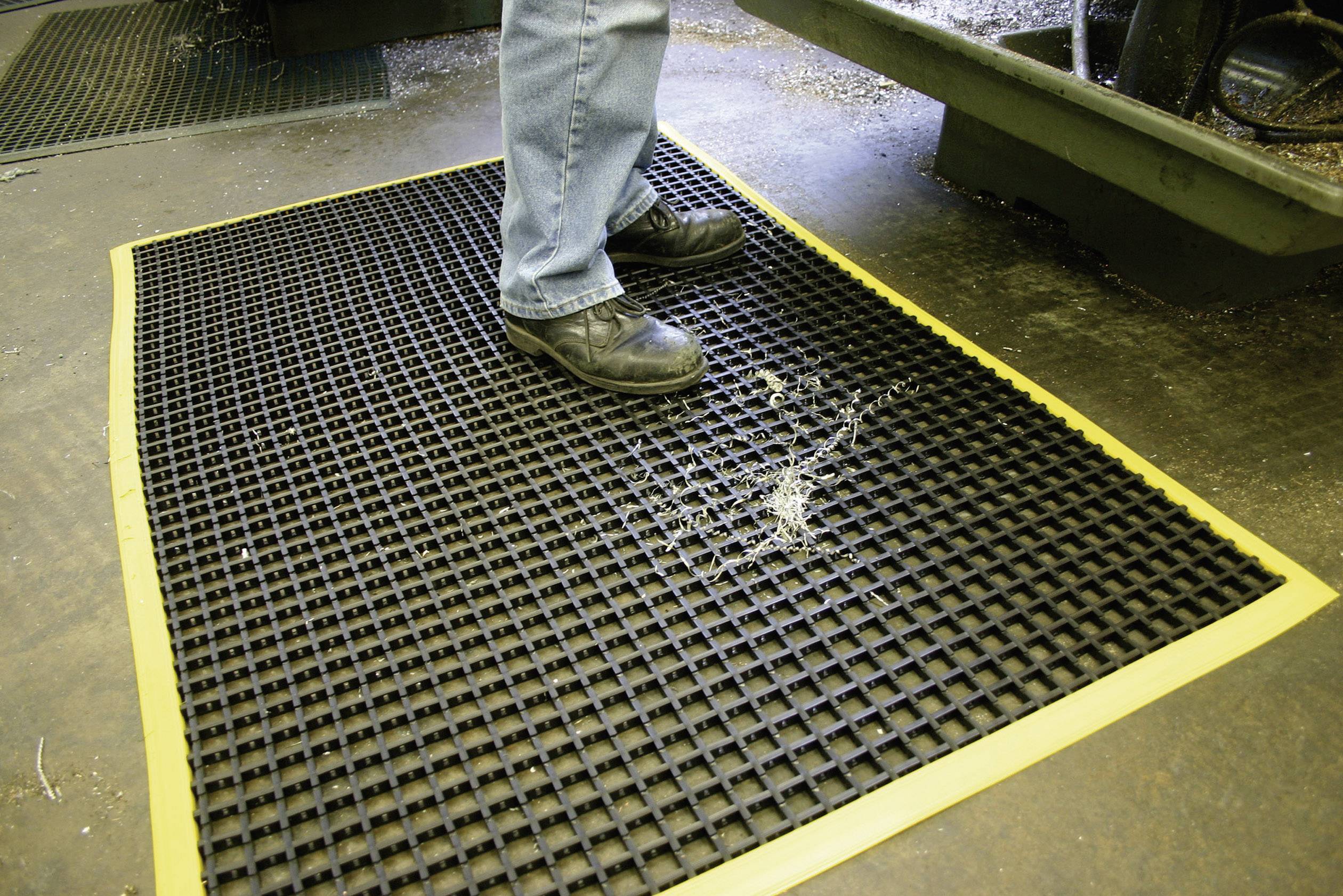 Une personne se tient sur un tapis de caoutchouc noir à motif de grille avec un bord jaune. Le tapis est posé sur un sol industriel.