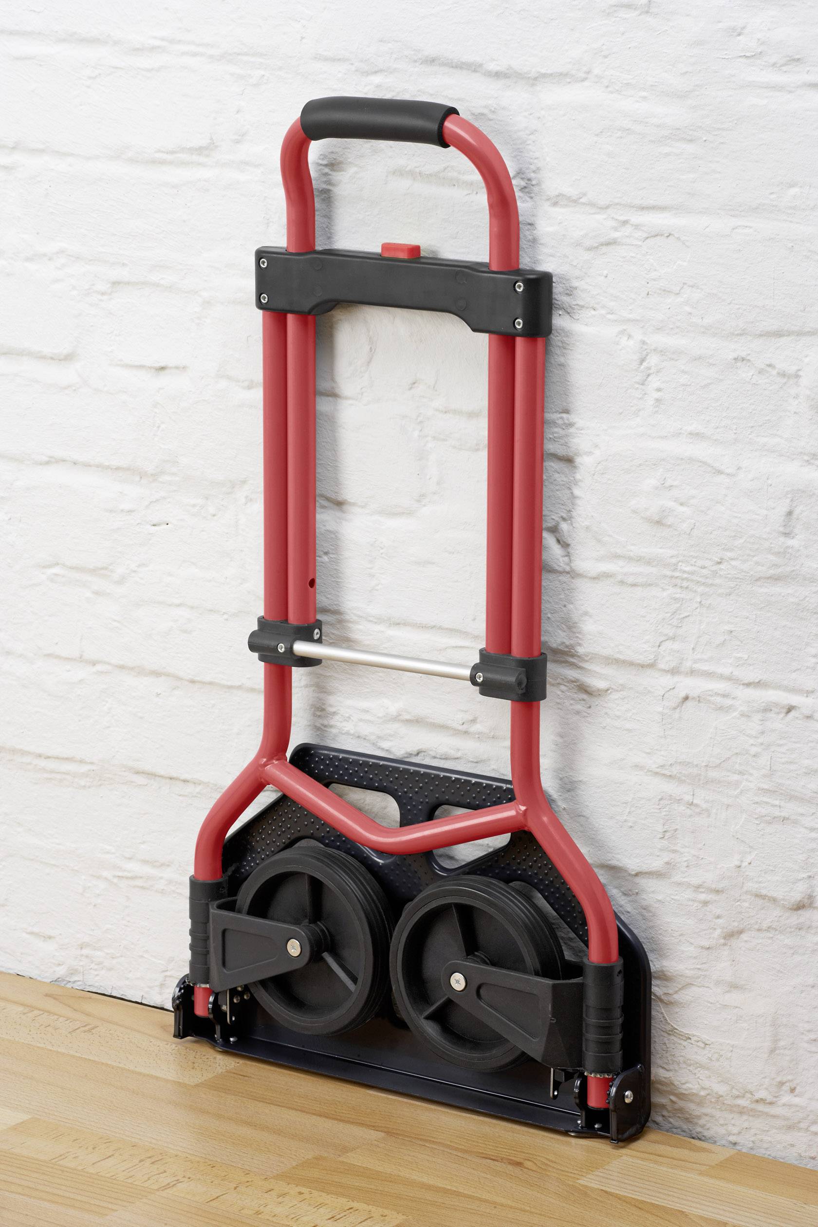 Une voiture pliante rouge avec des roues noires est appuyée contre un mur de briques blanches sur un plancher en bois.