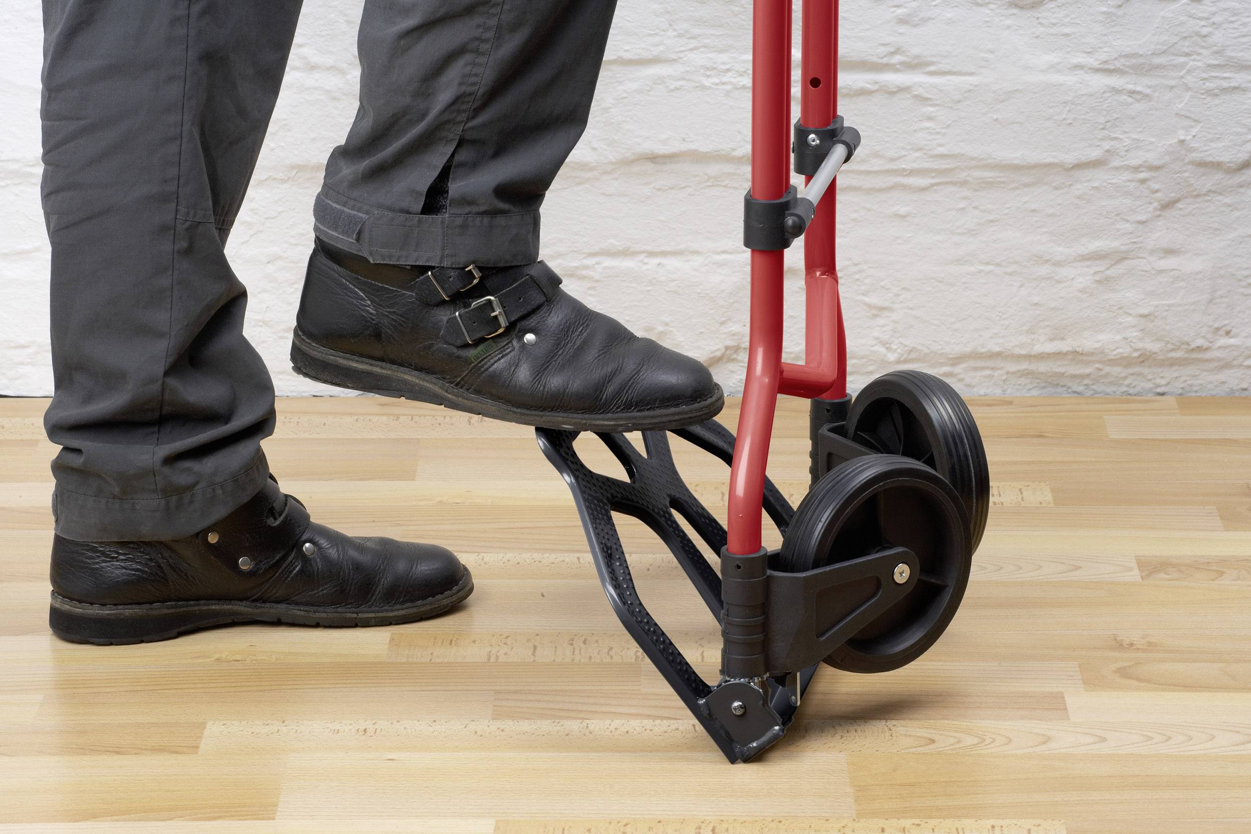 Une personne actionne un diable rouge avec son pied. Le diable est posé sur un plancher en bois devant un mur de briques blanches.
