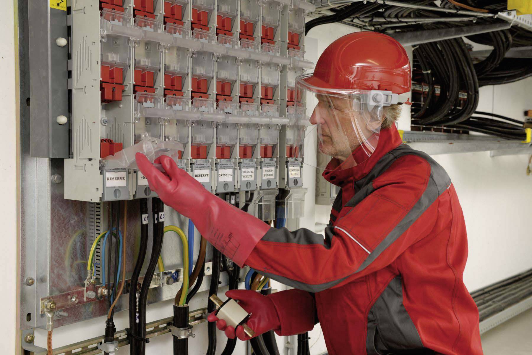 Un technicien en vêtements de protection rouge vérifie un tableau électrique dans un bâtiment. Il porte un casque et des lunettes de protection.
