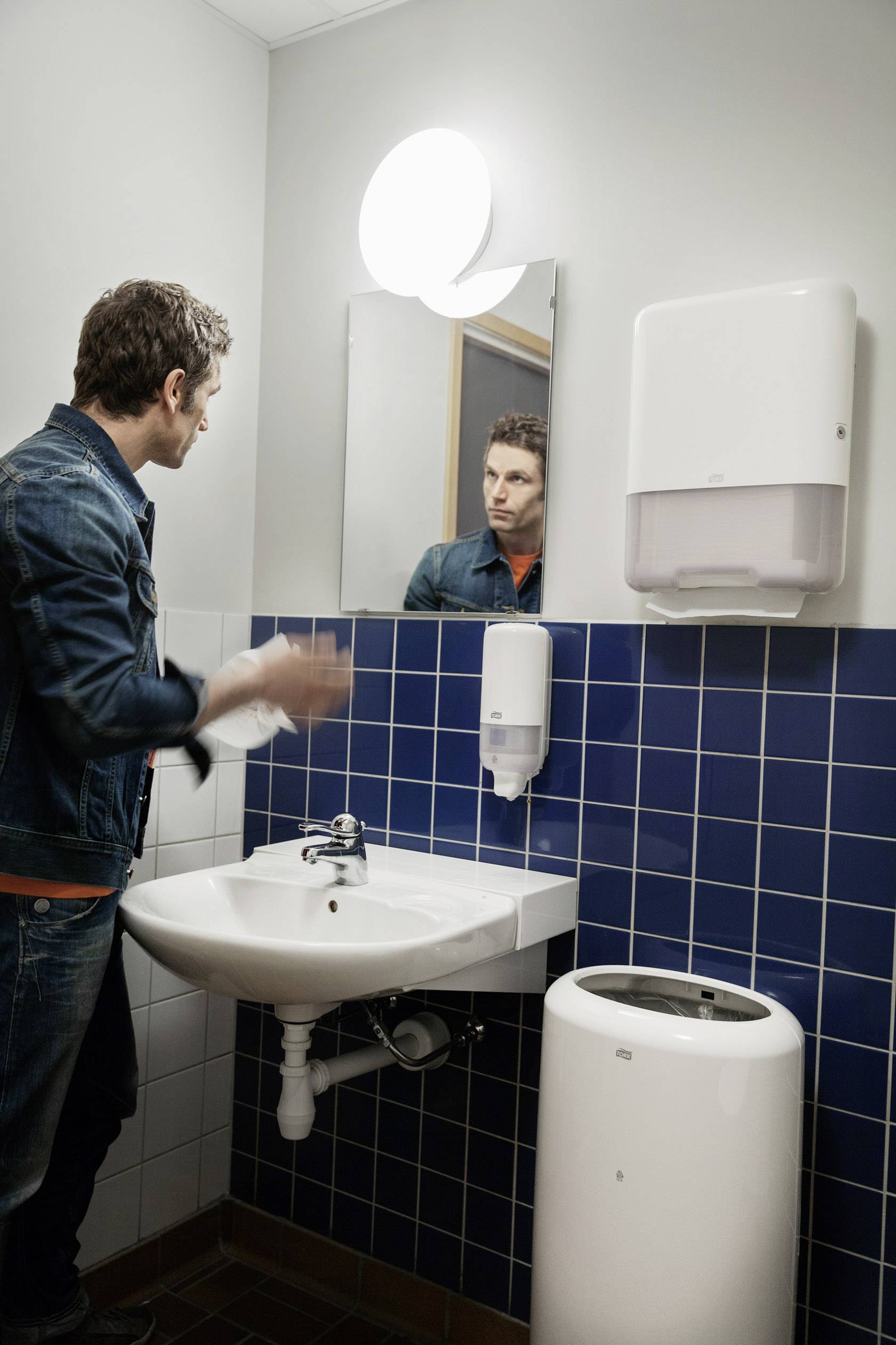 Un homme se tient dans une salle de bain devant un miroir. Il regarde son reflet. À côté de lui se trouvent un lavabo et un distributeur d'essuie-mains en papier.