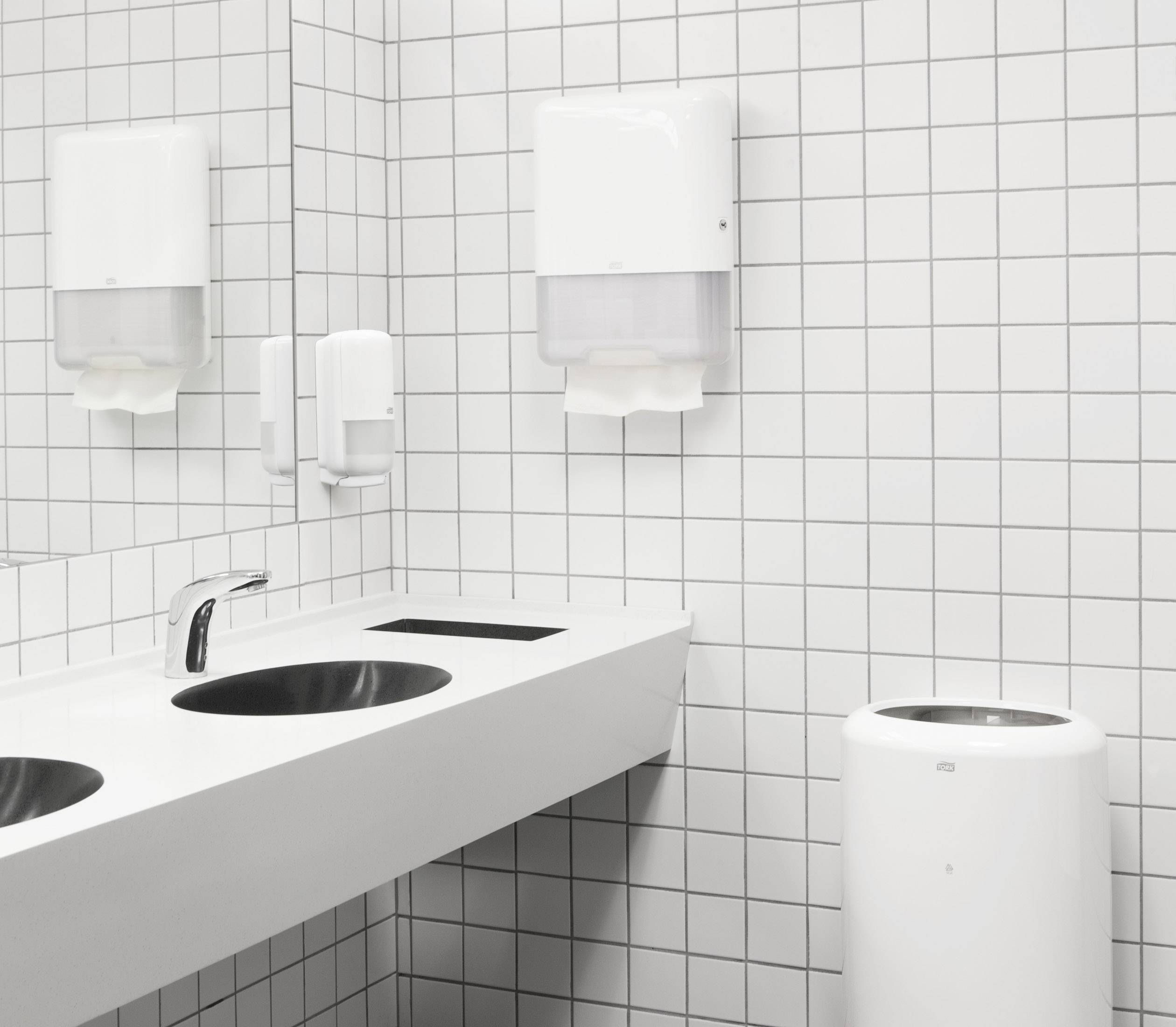 Une salle de bain moderne avec des murs carrelés de blanc, deux lavabos ronds, un distributeur de serviettes et une poubelle.