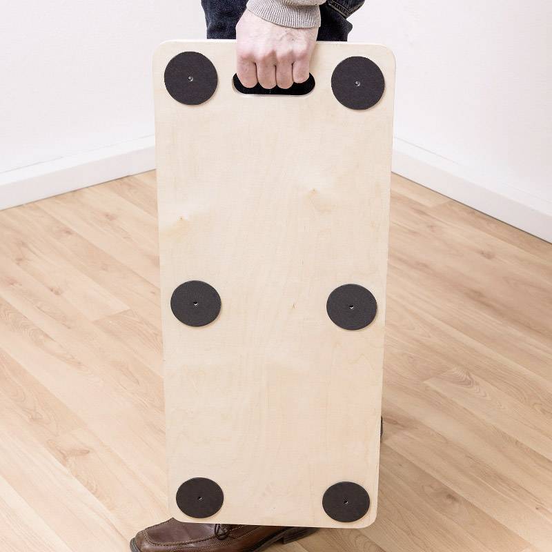 Une personne tient un plateau rectangulaire en bois avec une poignée, qui possède six pieds ronds noirs, au-dessus d'un plancher en bois.