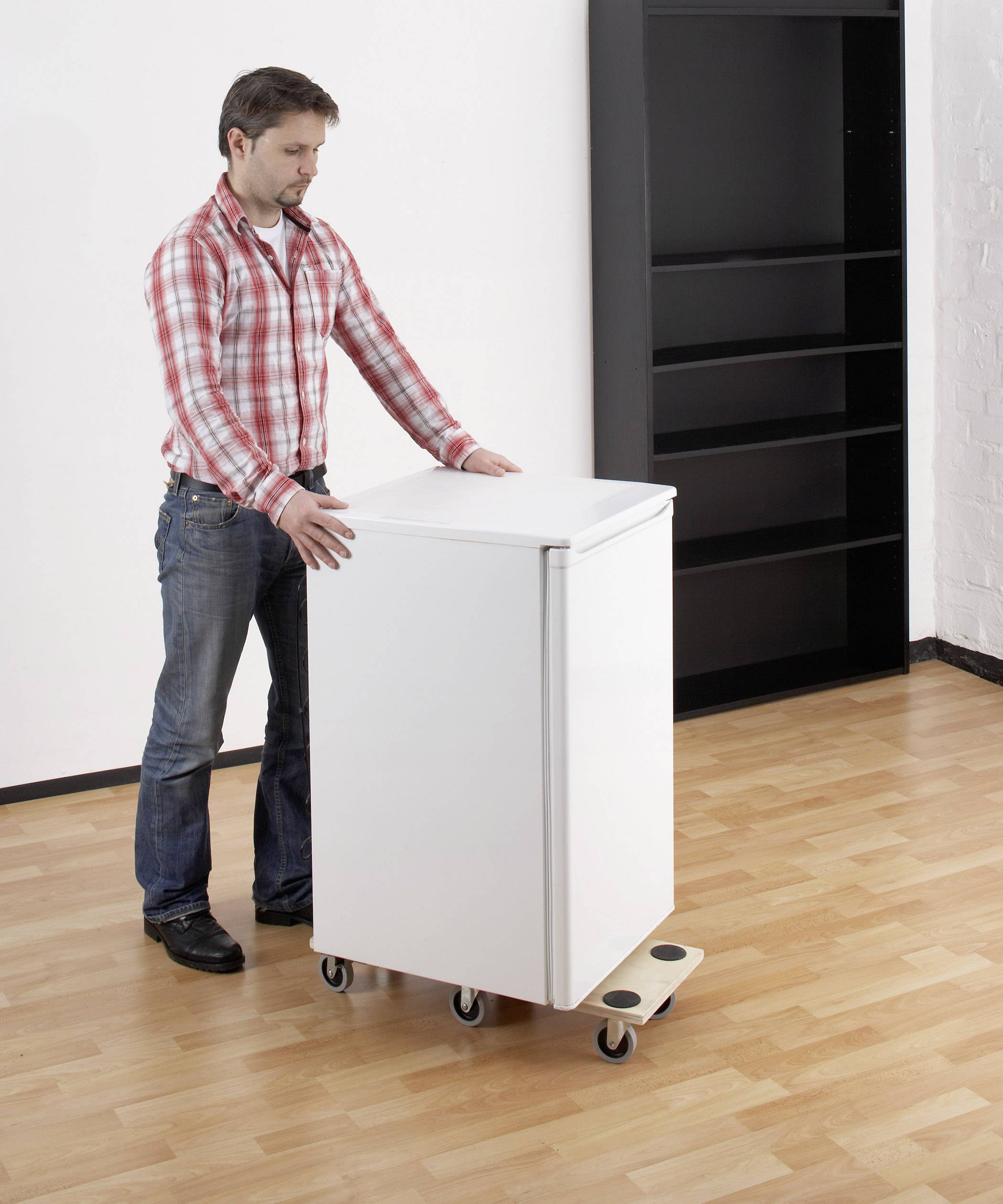 Un homme déplace difficilement une armoire sur un chariot à roulettes sur un sol en bois dans une pièce avec une étagère noire en arrière-plan.
