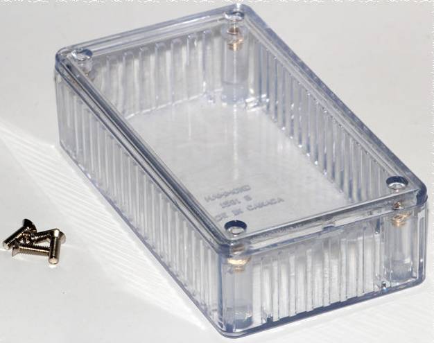 Boîte en plastique transparent au design strié, avec quatre vis à côté, sur un fond blanc.