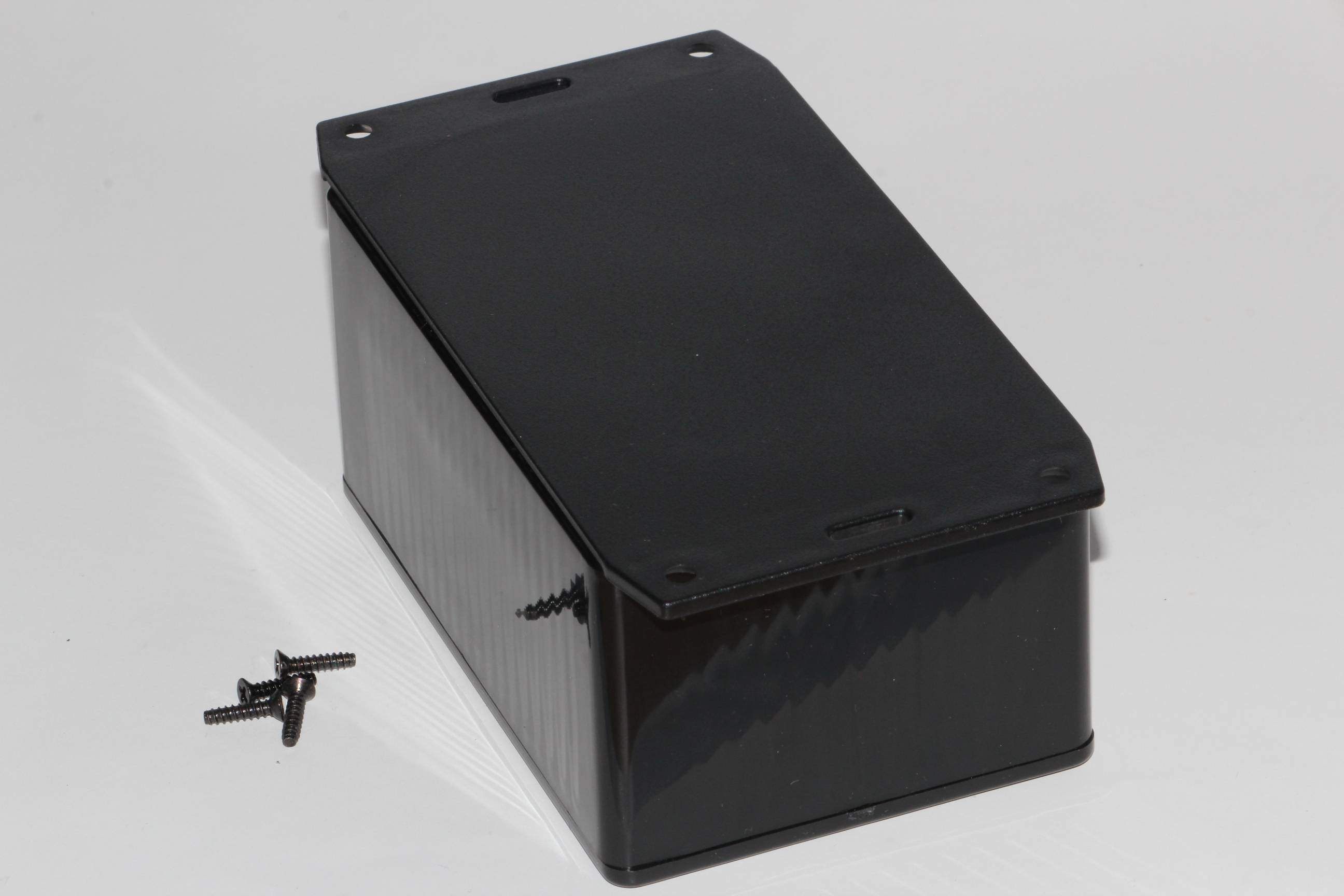 Boîtier rectangulaire noir avec un couvercle amovible, quatre vis noires sont posées à côté.