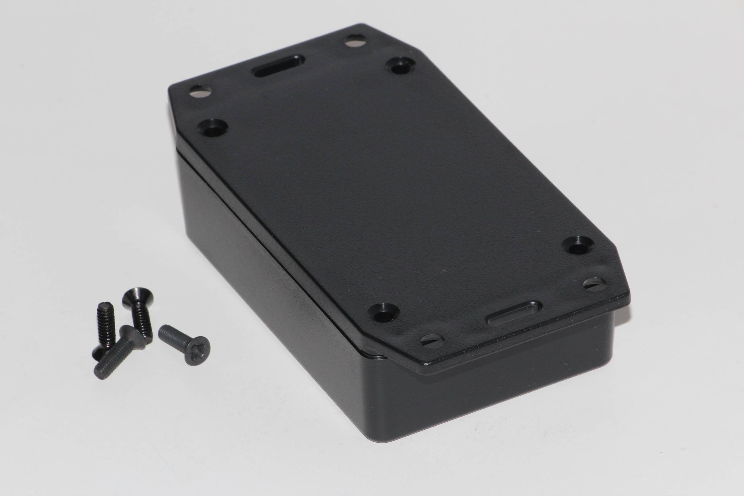 Un boîtier électronique rectangulaire noir avec quatre vis à côté, sur un fond blanc.