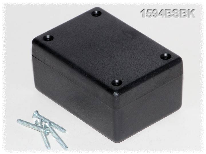 Boîtier noir rectangulaire aux angles arrondis. À côté se trouvent quatre vis argentées.