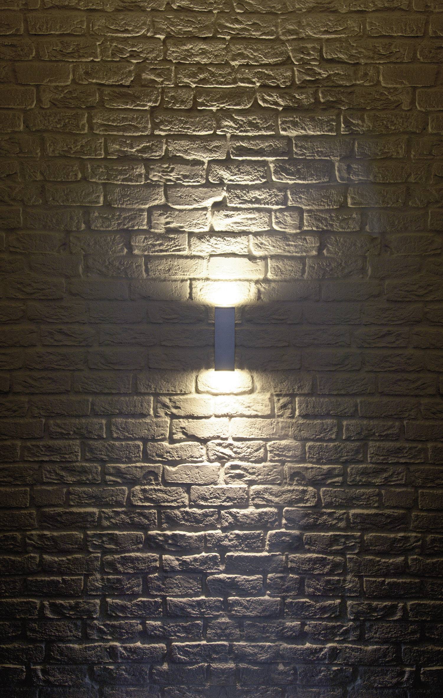 Applique murale sur un mur de briques, diffusant une lumière chaude vers le haut et le bas, mettant en valeur la texture des briques.