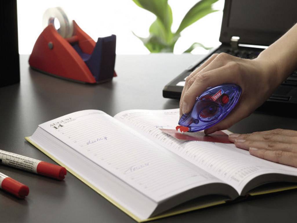 Une main utilise un correcteur à ruban sur un agenda ouvert. À l'arrière-plan, on aperçoit des stylos et un ordinateur portable.