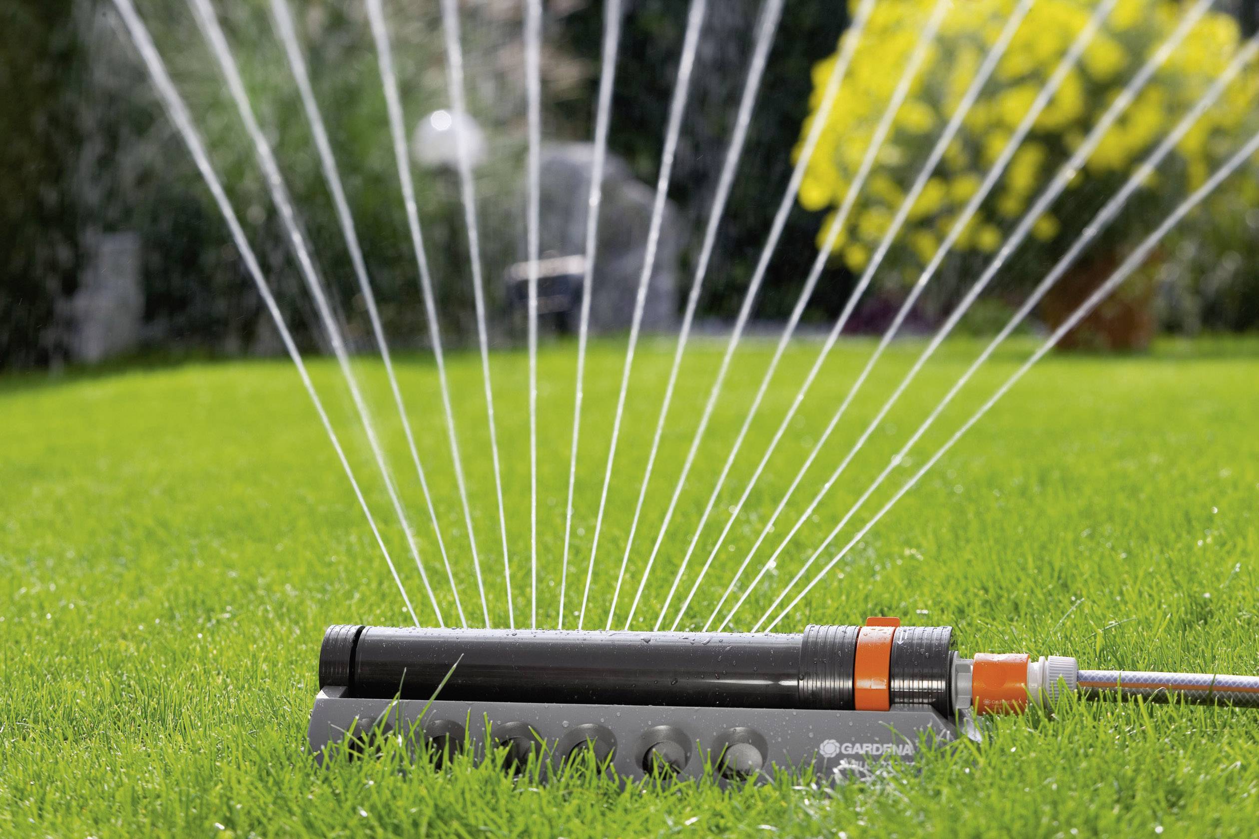 Un arroseur tournant irrigue uniformément une pelouse verte. Des arbustes et des arbres flous sont visibles en arrière-plan.