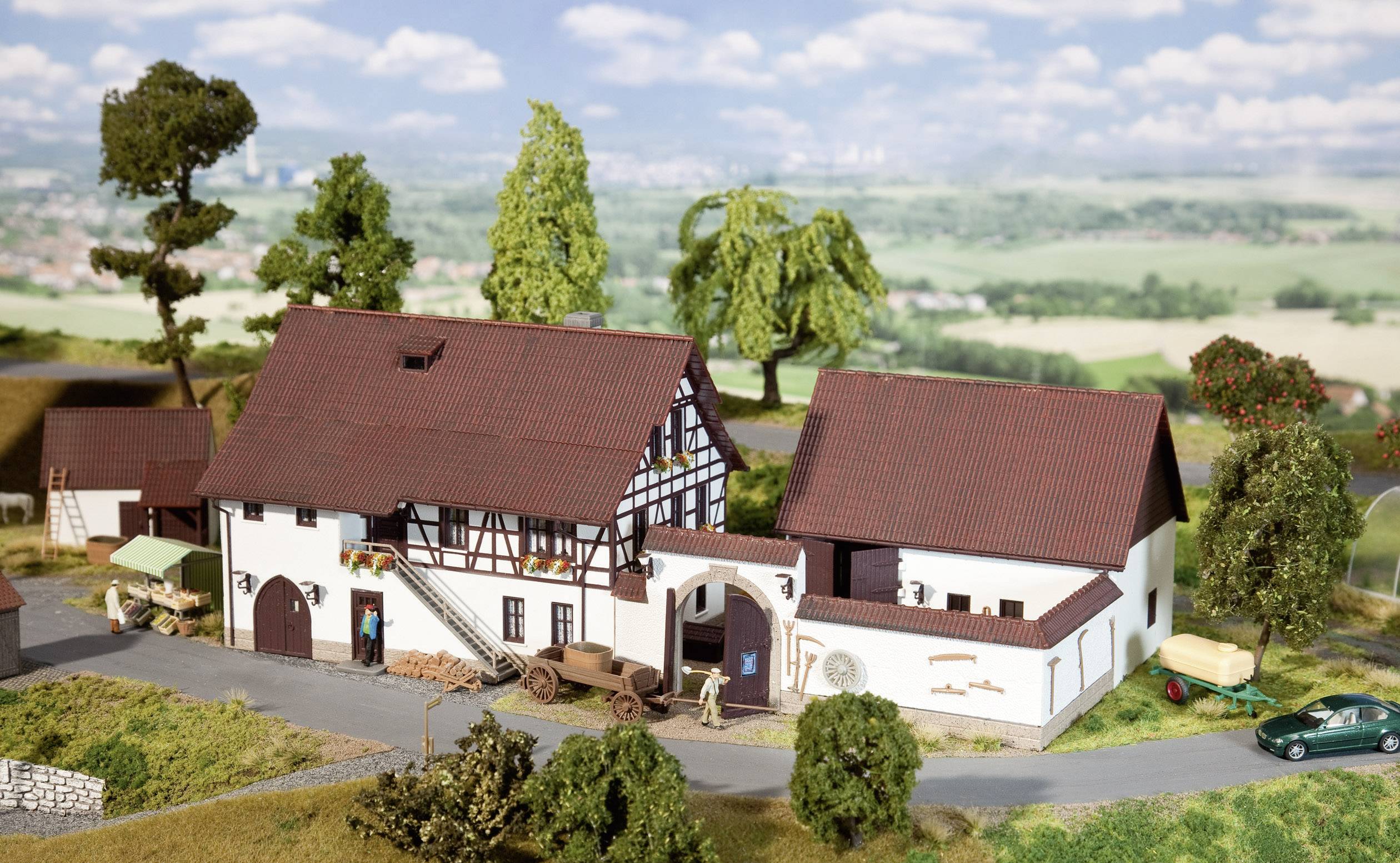 Maquette miniature d'une ferme avec une maison à colombages, des bâtiments annexes et des champs environnants. Représentation détaillée avec des arbres et des véhicules.