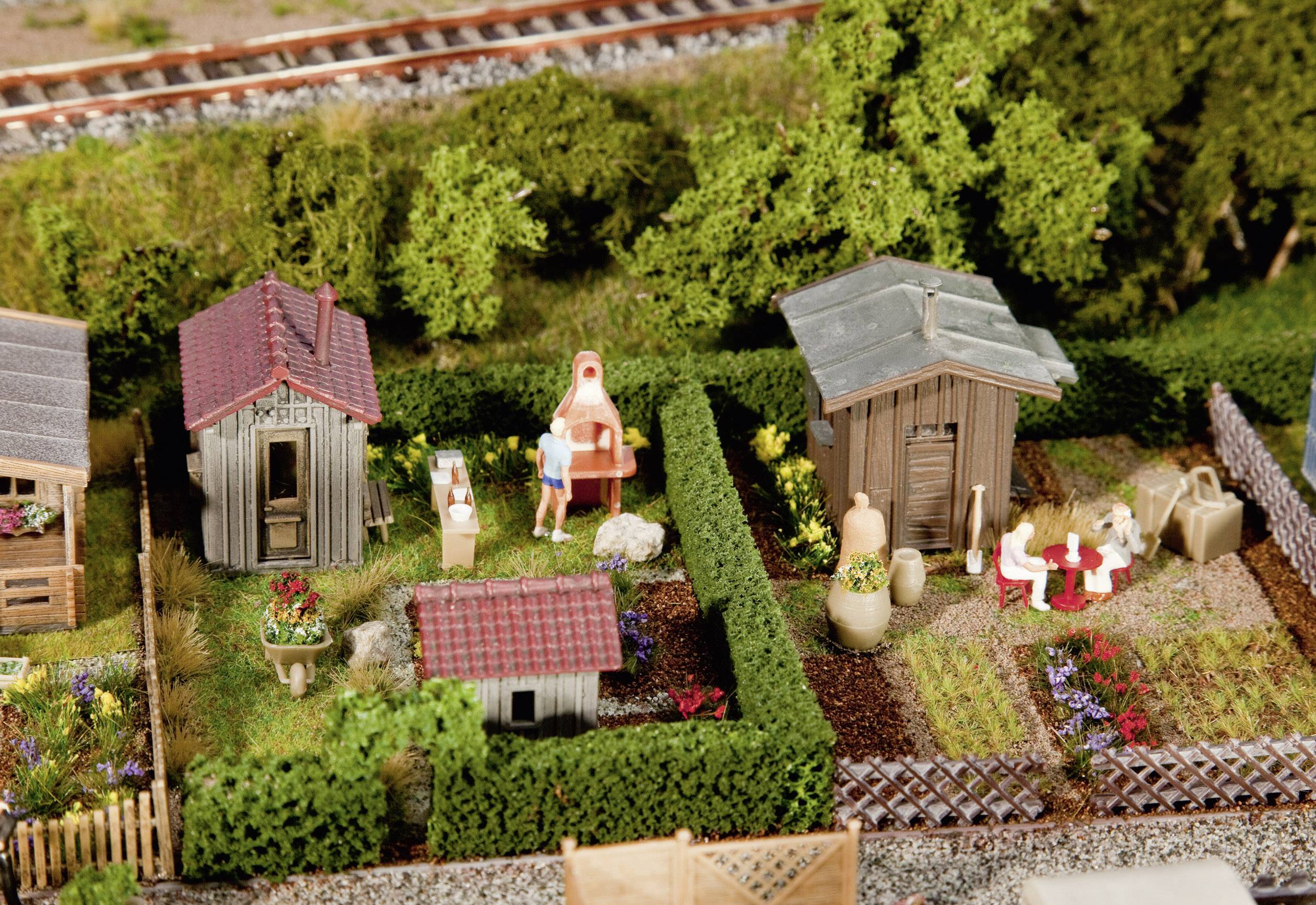 Modèle miniature d'un paysage de jardin avec deux petits bâtiments, des personnes faisant un barbecue et mangeant, entourées de plantes et de clôtures.