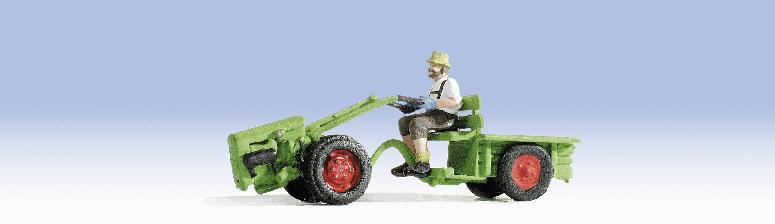 Un tracteur miniature avec un conducteur assis portant des vêtements traditionnels sur un fond bleu. Le tracteur est vert avec des roues rouges.