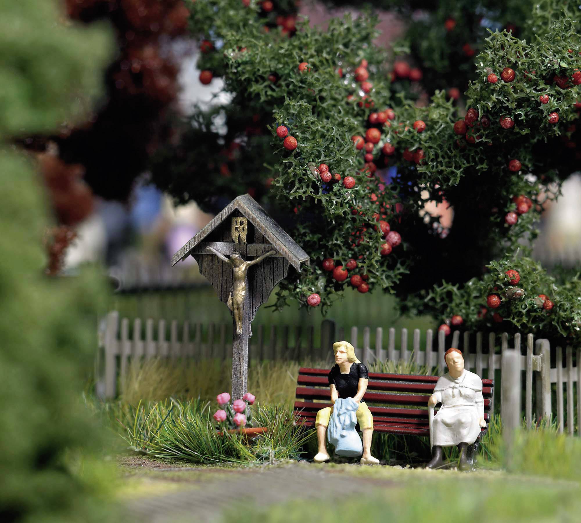 Une scène miniature montre des personnages assis sur un banc dans un parc, avec des arbustes en fleurs et un crucifix en arrière-plan.