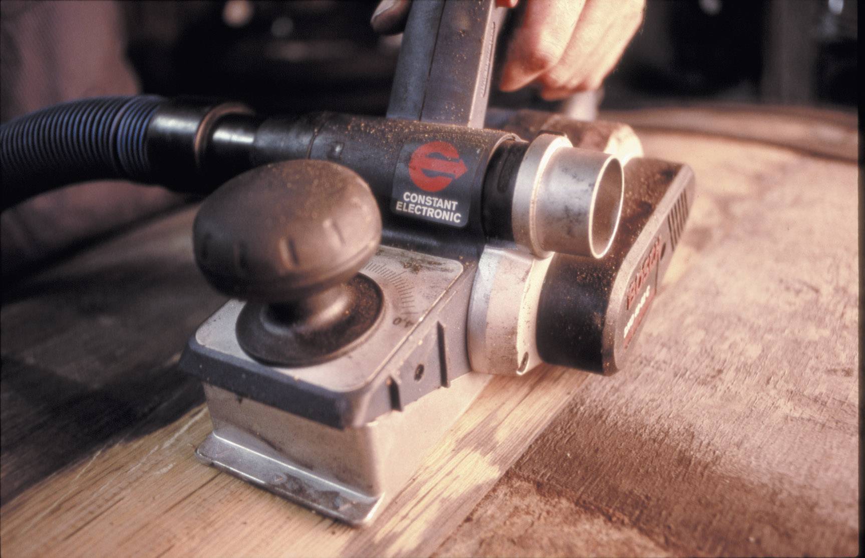 Une main utilise un rabot électrique pour lisser la surface d'une planche en bois. La poussière est aspirée par un tuyau.