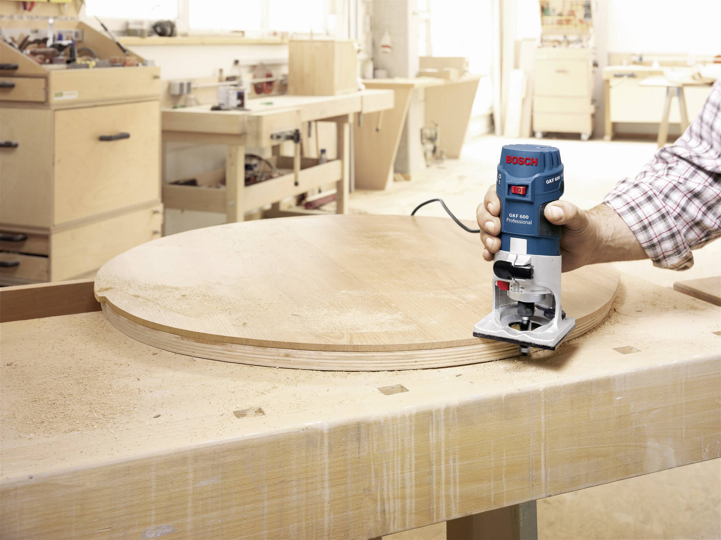 Un homme travaille une table ronde en bois dans un atelier à l'aide d'une défonceuse électrique. Des outils sont visibles en arrière-plan.