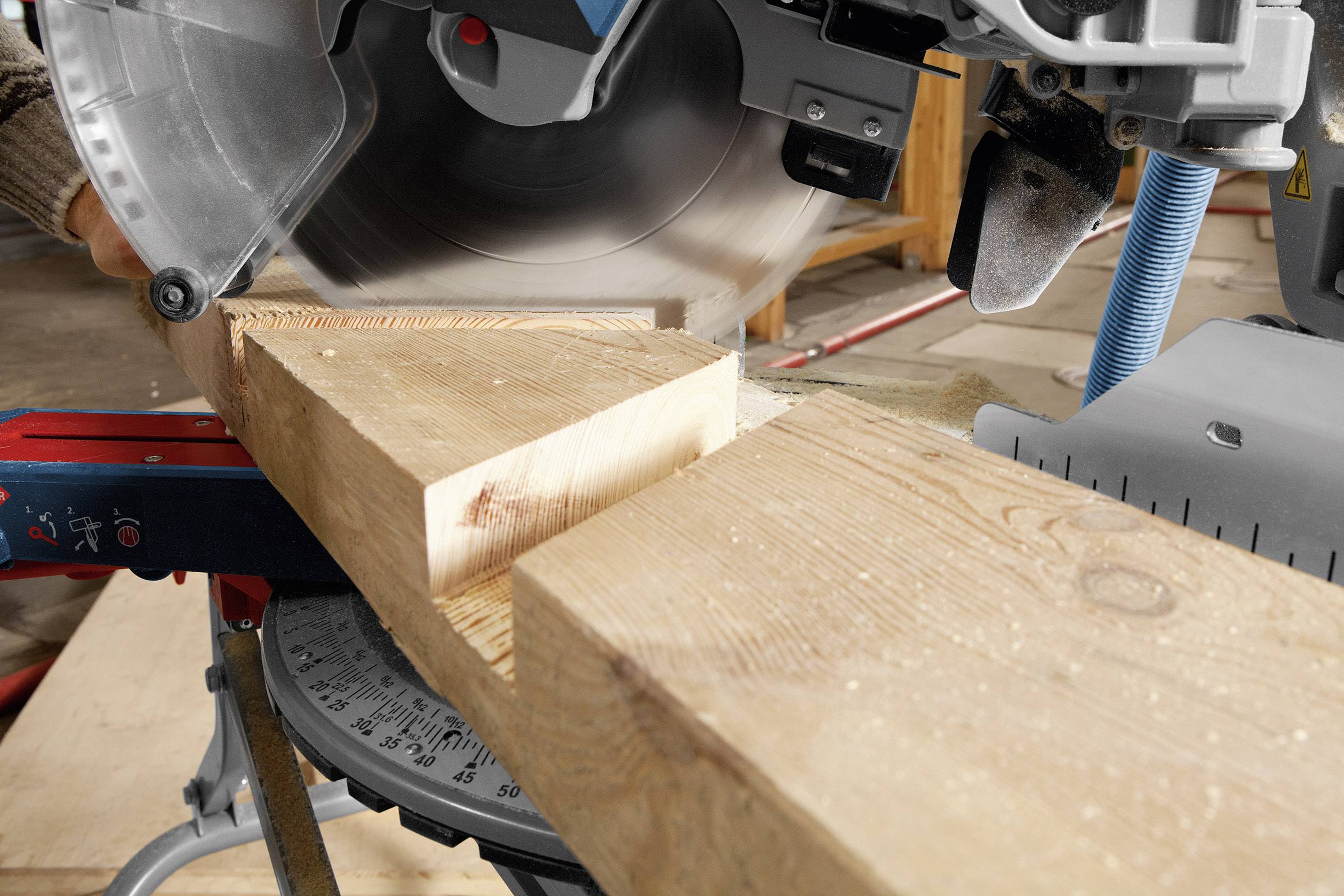 Une scie circulaire électrique coupe une planche de bois. La scie est en mouvement, des copeaux de bois volent. Un atelier est visible en arrière-plan.