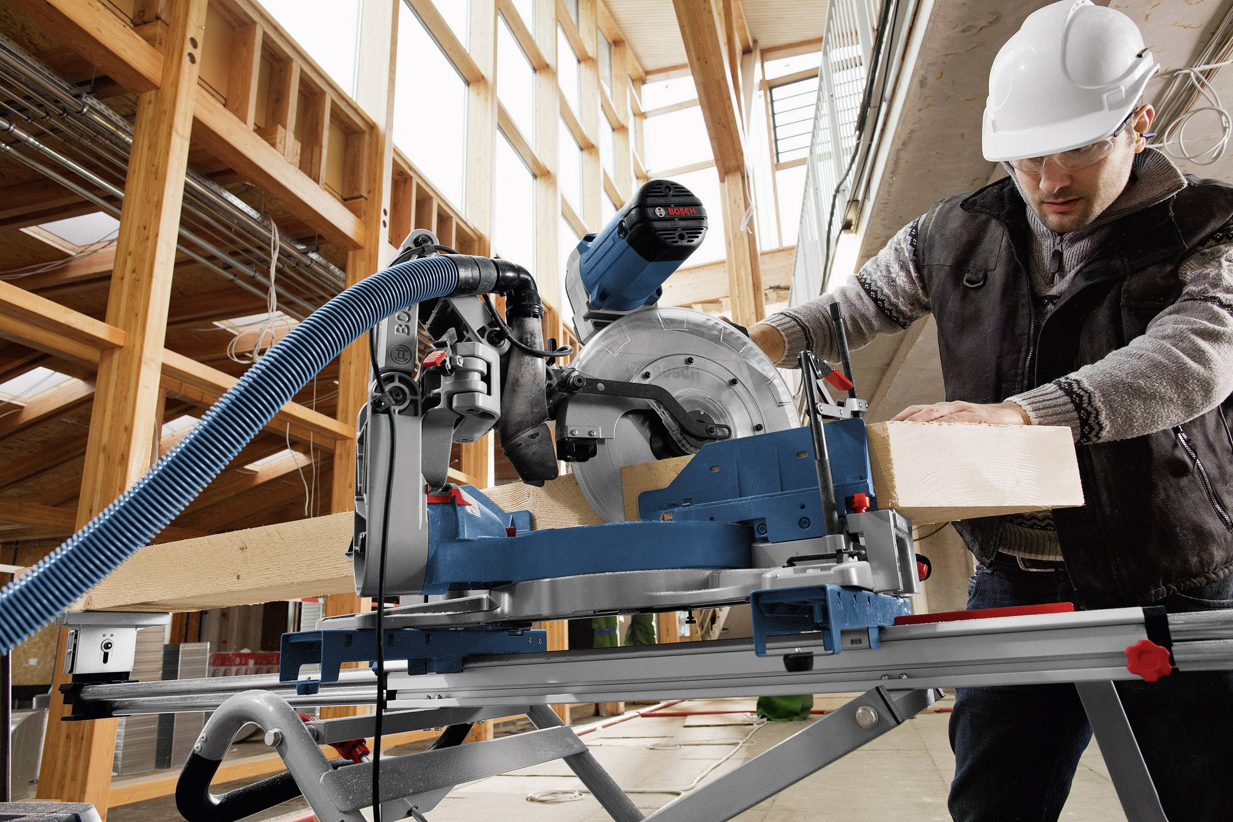 Ouvrier portant un casque de protection utilisant une scie circulaire dans un atelier. Une poutre en bois est en cours de découpe, avec des machines modernes et une structure en bois en arrière-plan.