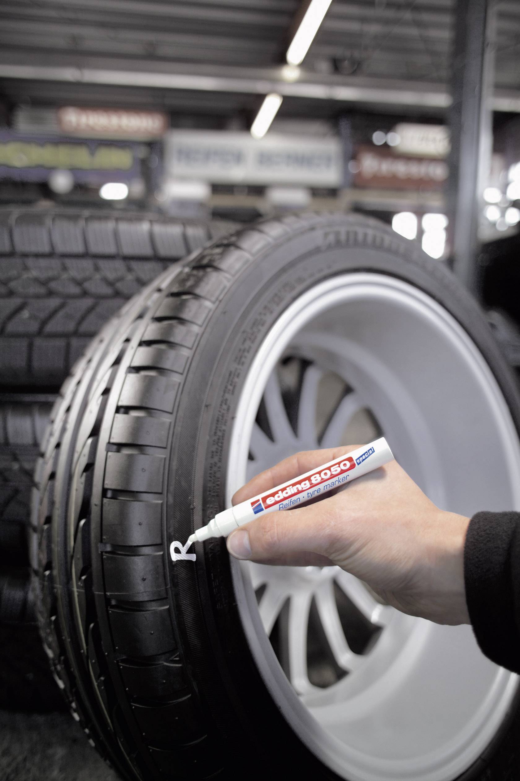 Une main marque un pneu de voiture avec un marqueur blanc. À l'arrière-plan, plusieurs pneus empilés sont visibles.