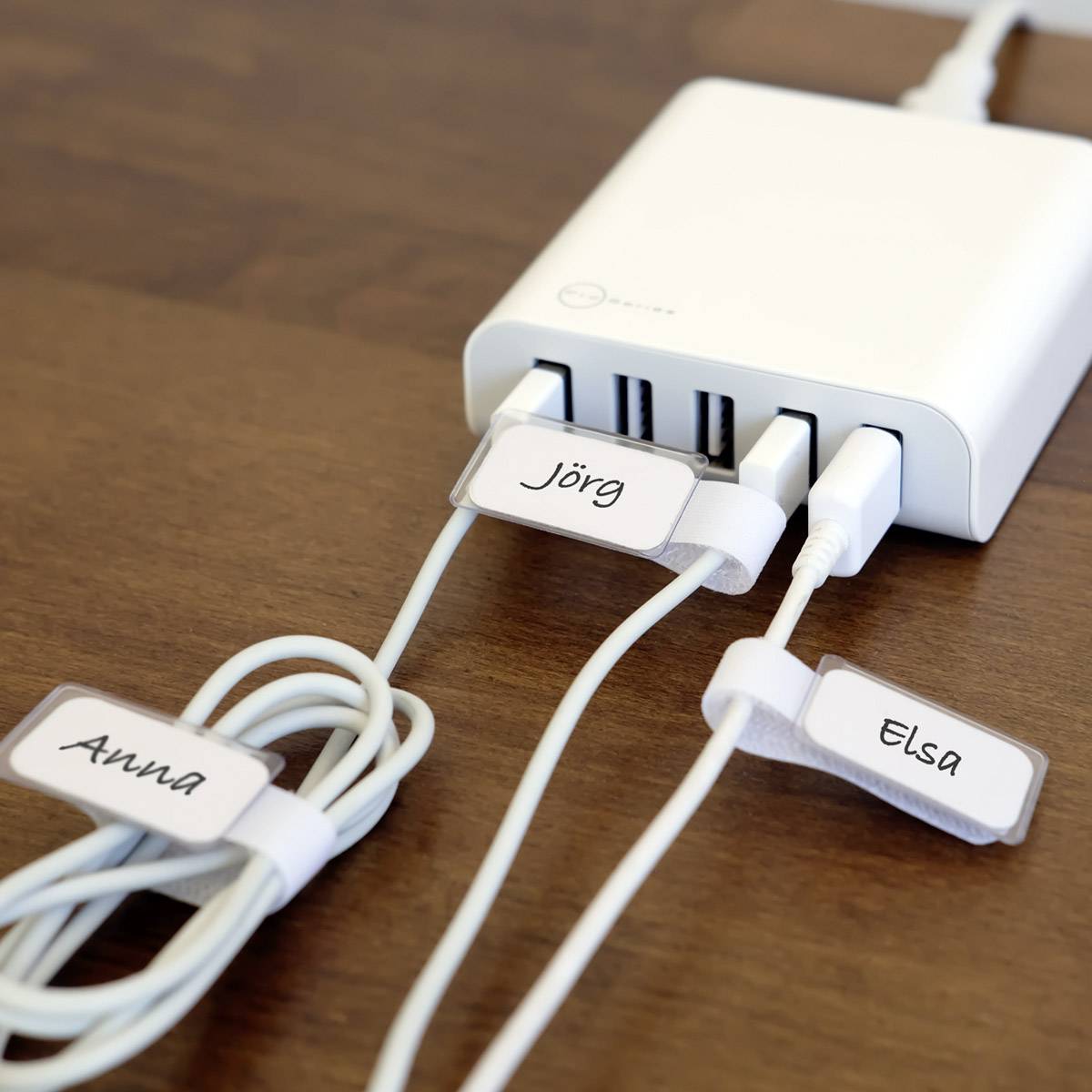 Trois câbles USB étiquetés ('Jörg', 'Anna', 'Elsa') sont branchés sur un hub USB blanc posé sur une table en bois.