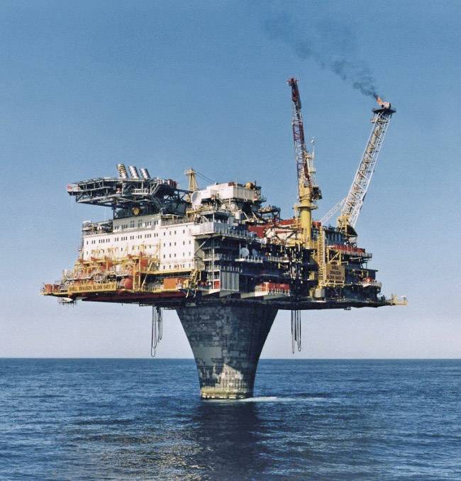 Une grande plate-forme de forage offshore se dresse isolée en pleine mer, entourée de ciel et d'eau bleus, avec ses grues et ses structures de travail.