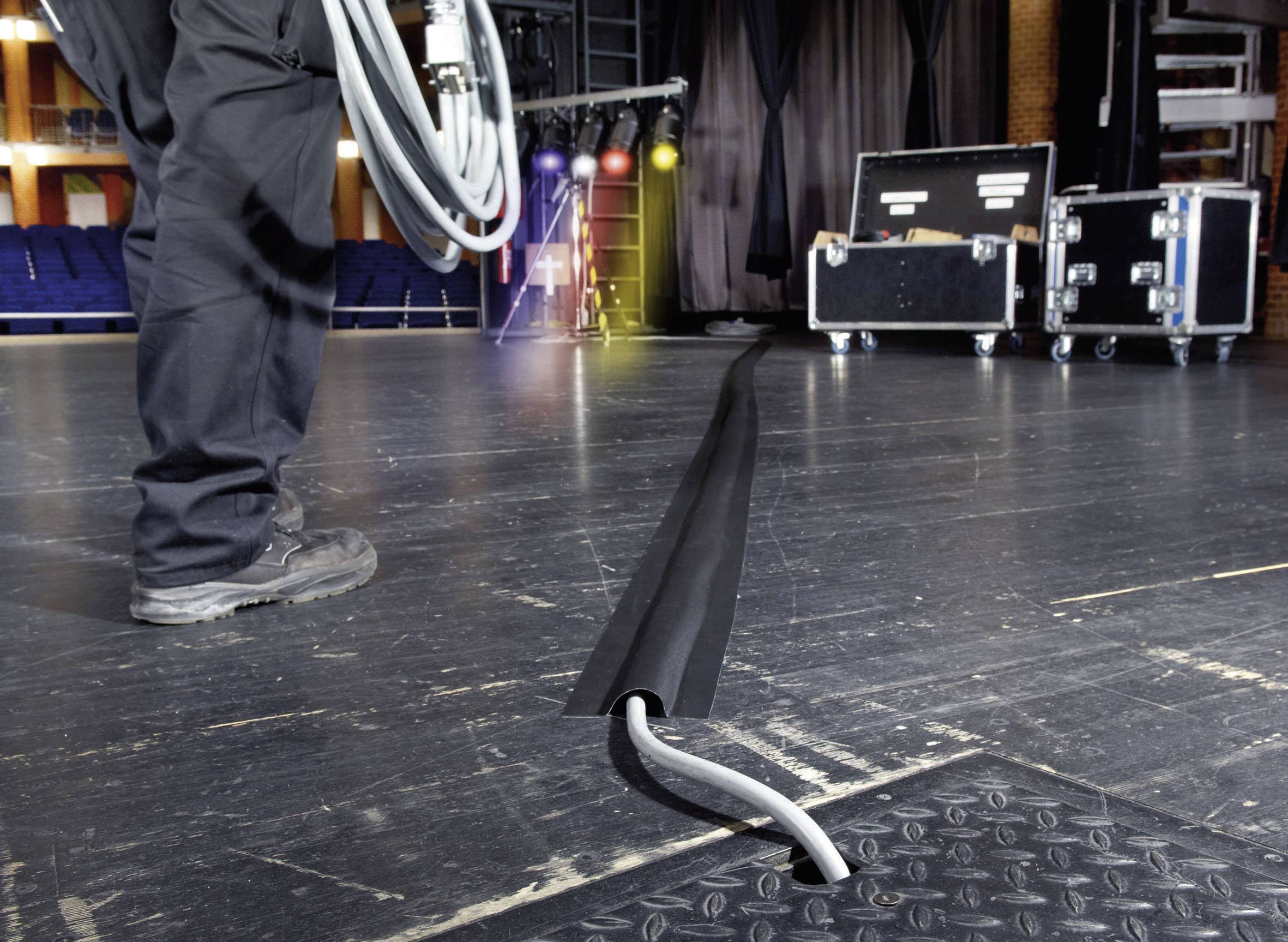 Une personne sur une scène installe un câble sous une gaine de protection. Des projecteurs et des caisses de transport sont visibles en arrière-plan.