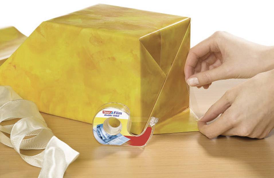 Une personne emballe un cadeau avec du papier jaune et du ruban adhésif. Un nœud et un rouleau de ruban adhésif sont posés sur la table.