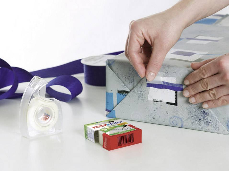 Une personne emballe un cadeau avec un ruban bleu et du ruban adhésif. Sur la table, on peut voir du ruban adhésif et un emballage de film.