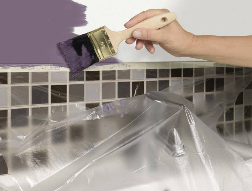 Une main peint un mur carrelé avec un pinceau de couleur lilas. Les carreaux sont recouverts de film plastique.
