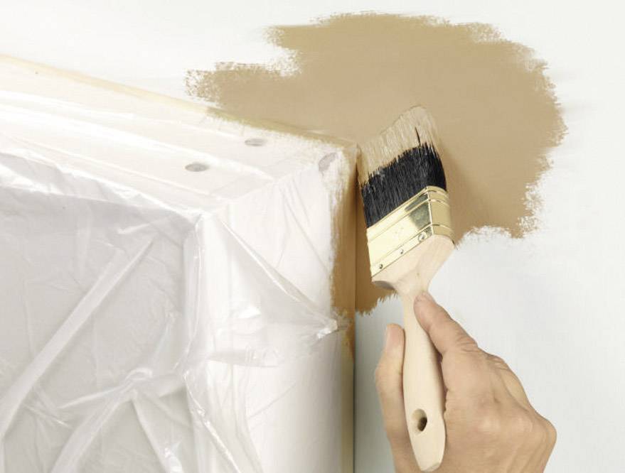 Une main peint un mur en marron avec un pinceau, tandis qu'un meuble est recouvert de film plastique.
