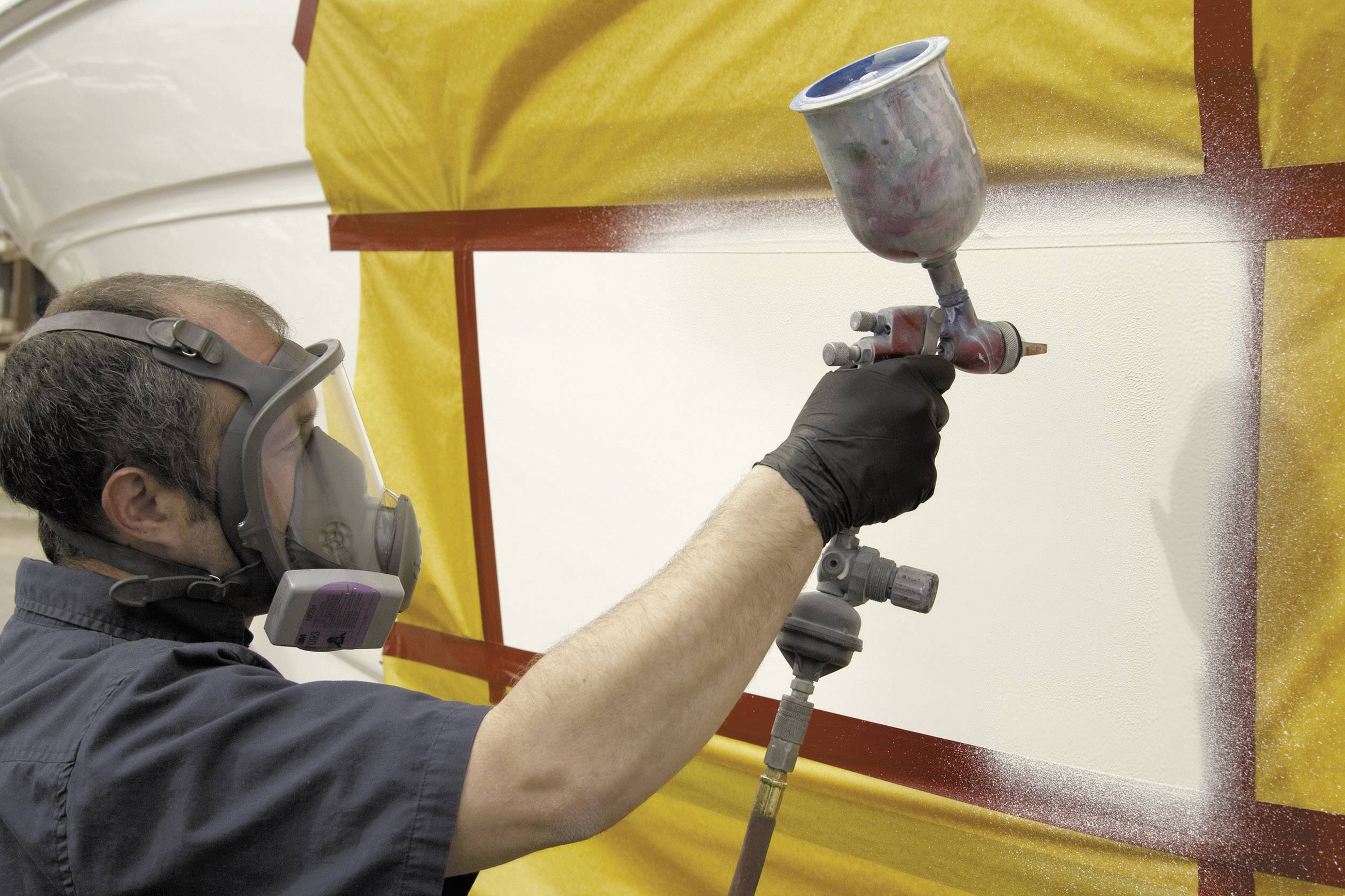 'Un homme porte un masque de protection et peint une surface avec un pistolet de pulvérisation. La zone est recouverte de film plastique.'