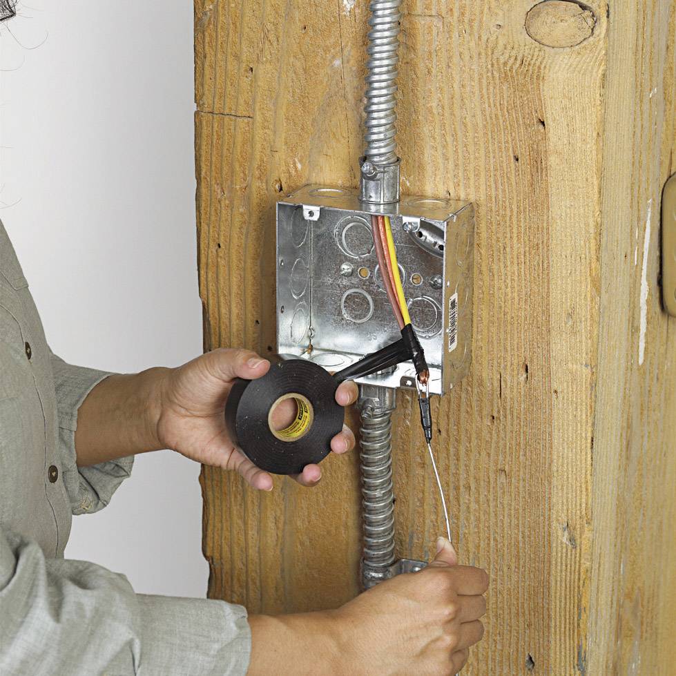 Une personne installe un câble électrique dans un boîtier métallique sur un mur, en utilisant du ruban adhésif noir pour le fixer.