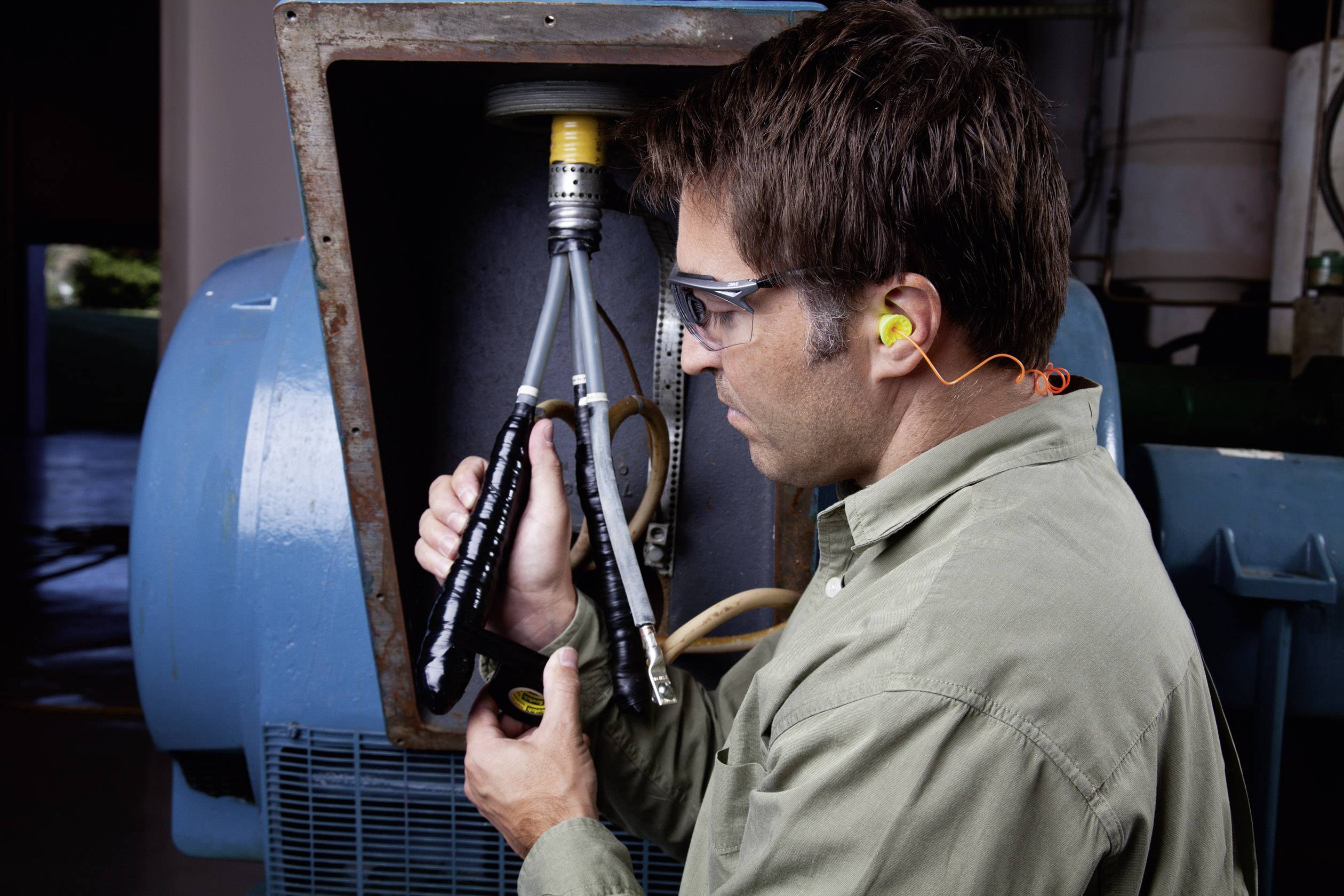 Un homme portant des lunettes de protection et des bouchons d'oreilles vérifie les connexions de câbles sur une grande machine industrielle à l'intérieur.