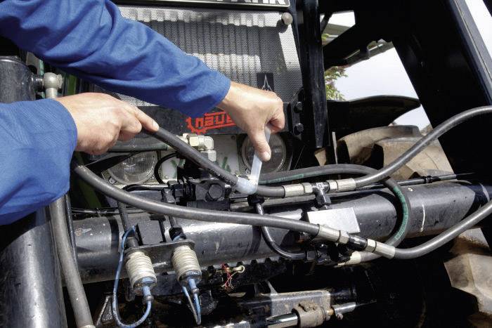 Une personne répare ou entretient les tuyaux hydrauliques d'un tracteur. Les mains tiennent des outils et des tuyaux sur un dispositif métallique.