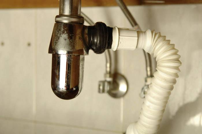 Un tuyau de siphon chromé sous un lavabo, relié à un tuyau de vidange blanc et flexible. Arrière-plan : mur carrelé.