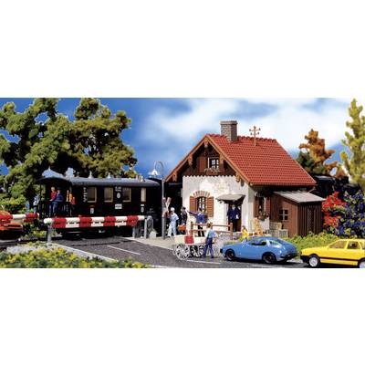 Une scène de train miniature avec un train noir à un passage à niveau, entouré de personnes, d'une maison, de voitures et d'arbres.