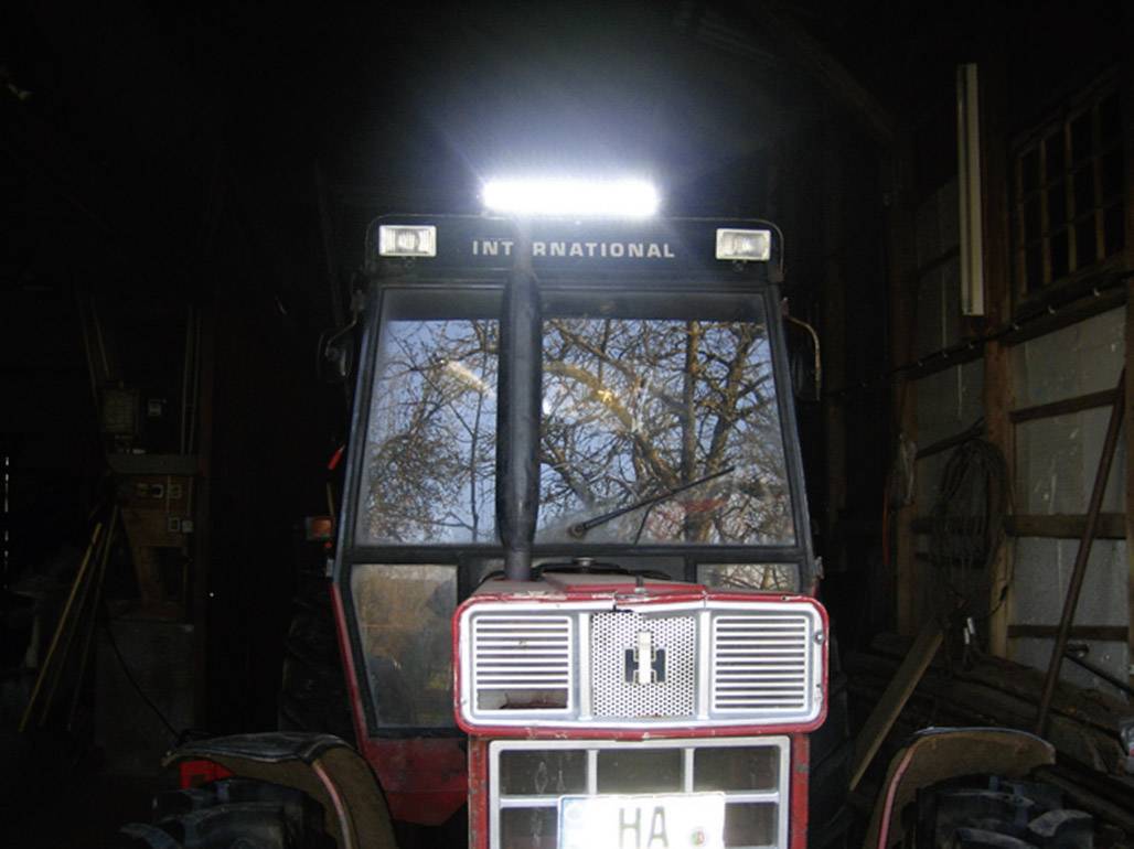Un tracteur est stationné dans une grange sombre, éclairé par un projecteur puissant sur le toit. Structure en bois en arrière-plan.