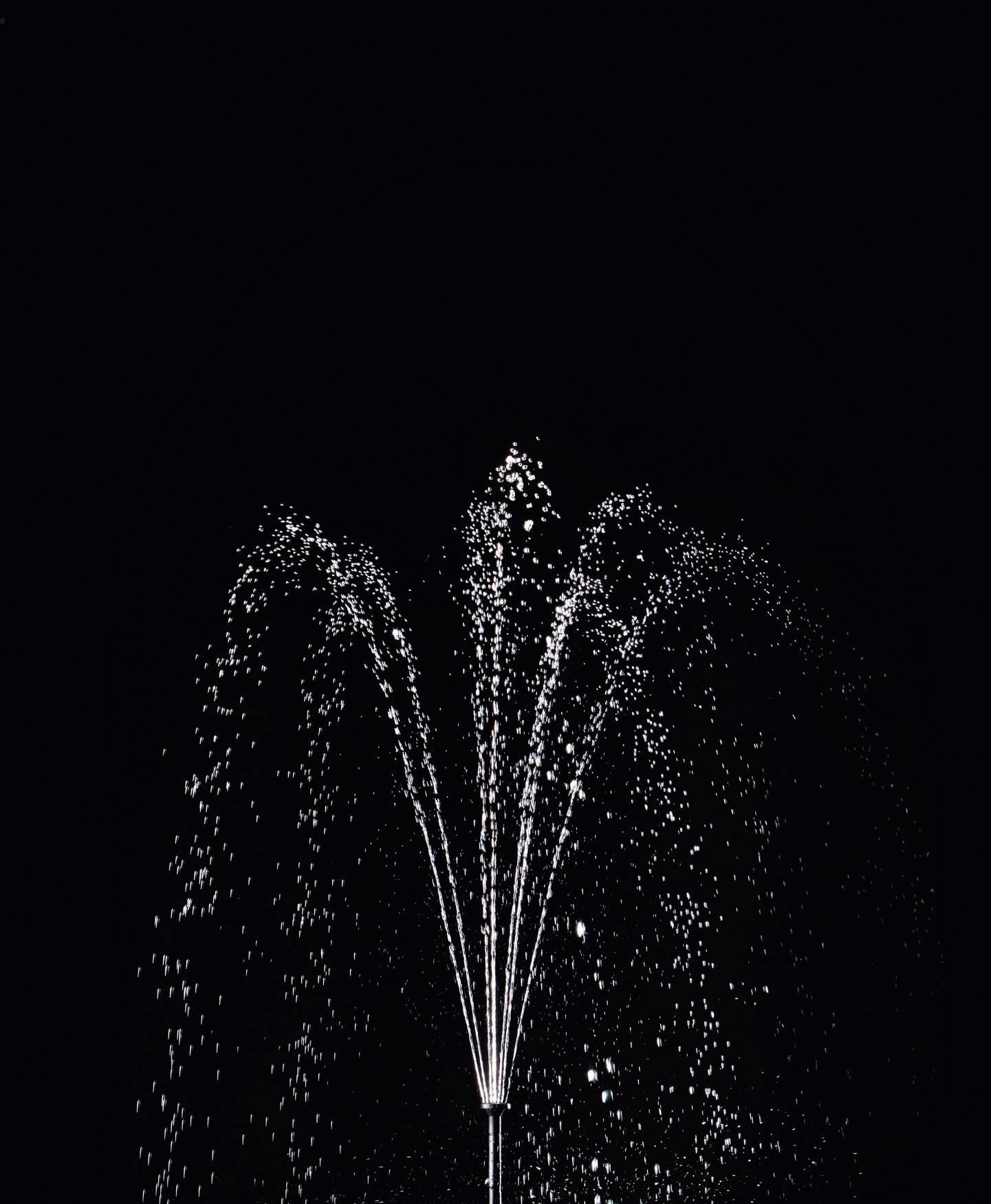 Une fontaine illuminée la nuit projette des jets d'eau jaillissant en arc devant un fond sombre.