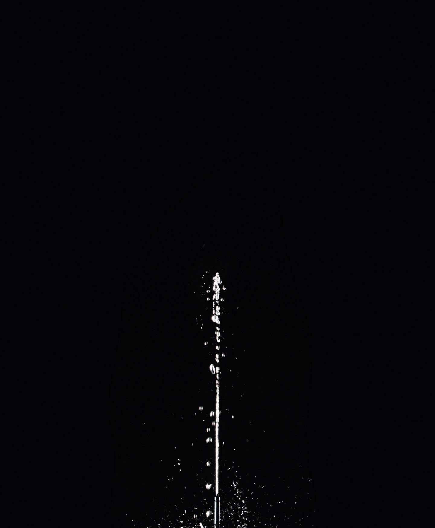 Un jet d'eau jaillit verticalement dans les airs devant un fond noir.