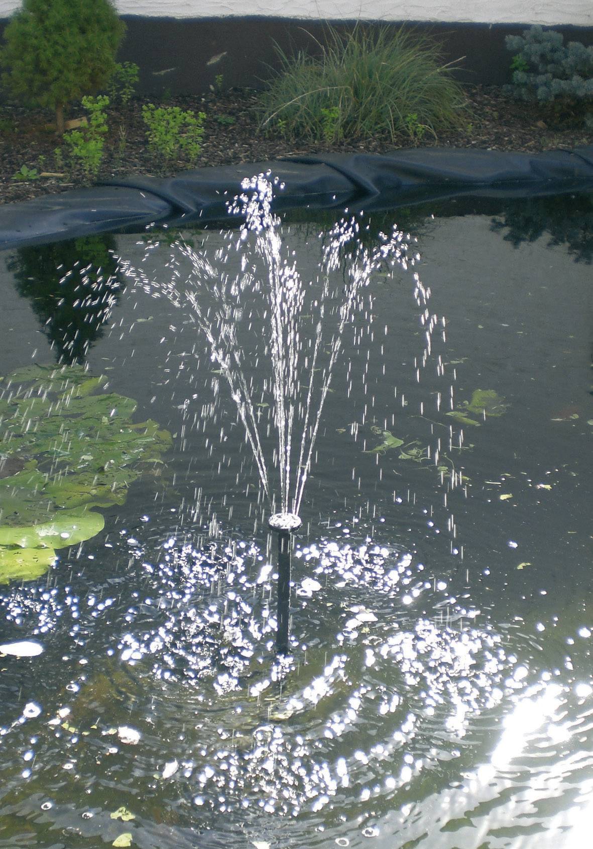 Une fontaine de jardin projette de l'eau en arc dans un bassin, entourée de nénuphars et de feuillage vert. La lumière du soleil se reflète à la surface de l'eau.