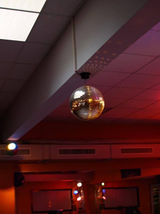 Une boule à facettes est suspendue au plafond d'une pièce éclairée en rouge, projetant des points lumineux sur les murs. Ambiance de fête et de danse.