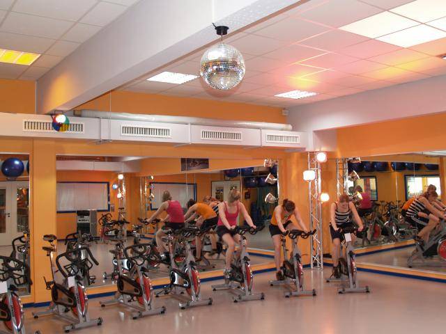 Des personnes s'entraînent sur des vélos fixes dans une salle de fitness aux murs recouverts de miroirs et éclairée comme une discothèque.